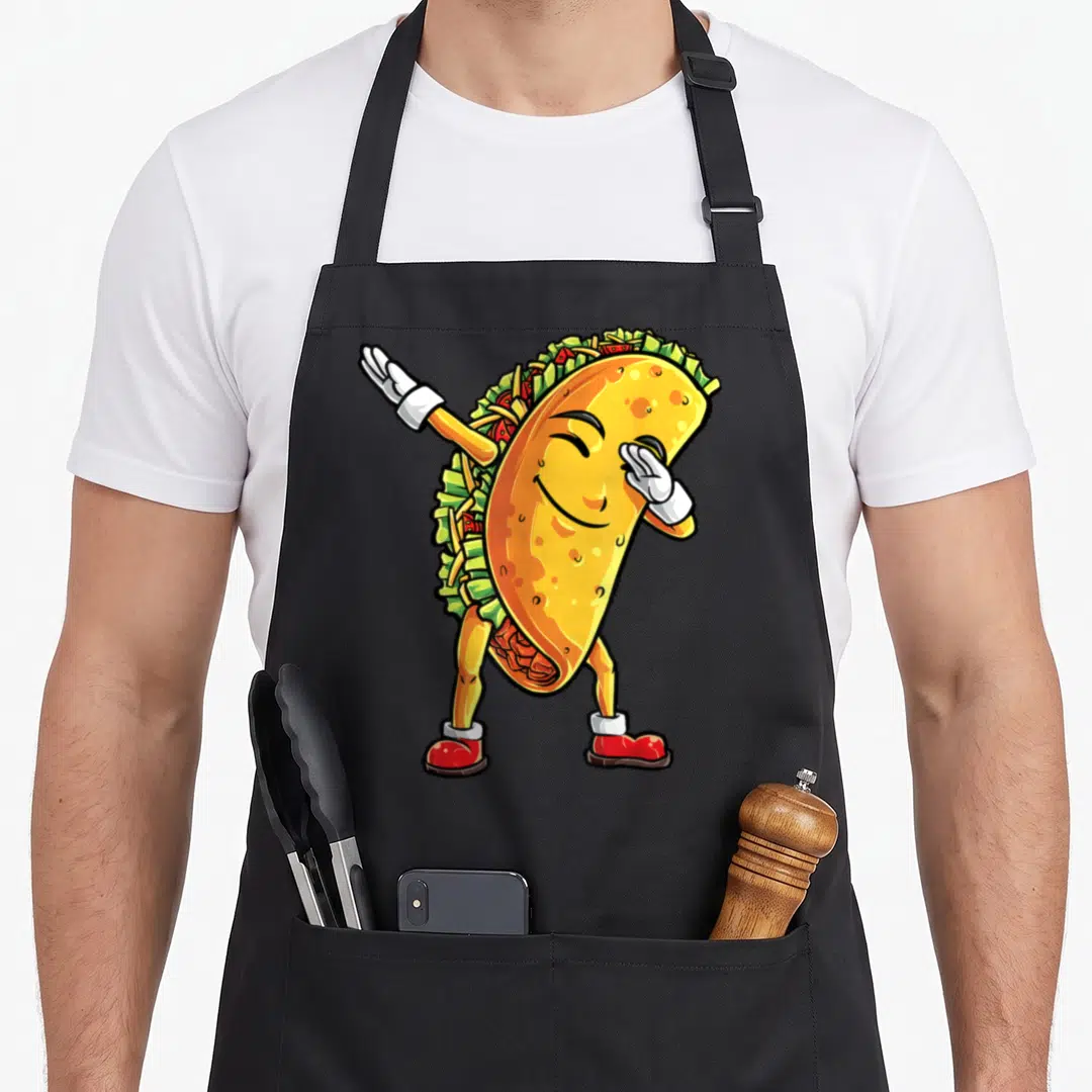 Taco mexican apron - Cartoon taco character dabbing in yellow green and red colors — shown on a black cotton canvas apron against a white background
