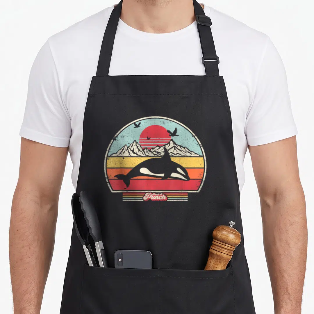 Funny killer orca apron - Retro grey orca swimming under red sun and mountains with Pack A Punch text — shown on a black cotton canvas apron against a white background