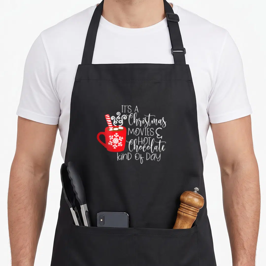 Hot women's christmas apron - Red mug with snowflake and candy cane next to white stacked text about Christmas movies — shown on a black cotton canvas apron against a white background