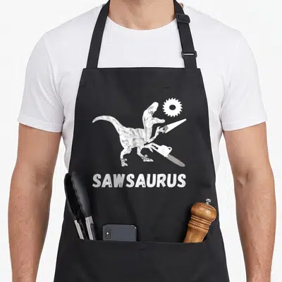 Dinosaur carpentry apron - White cartoon dinosaur holding chainsaw and hand saw with Sawsaurus text below — shown on a black cotton canvas apron against a white background