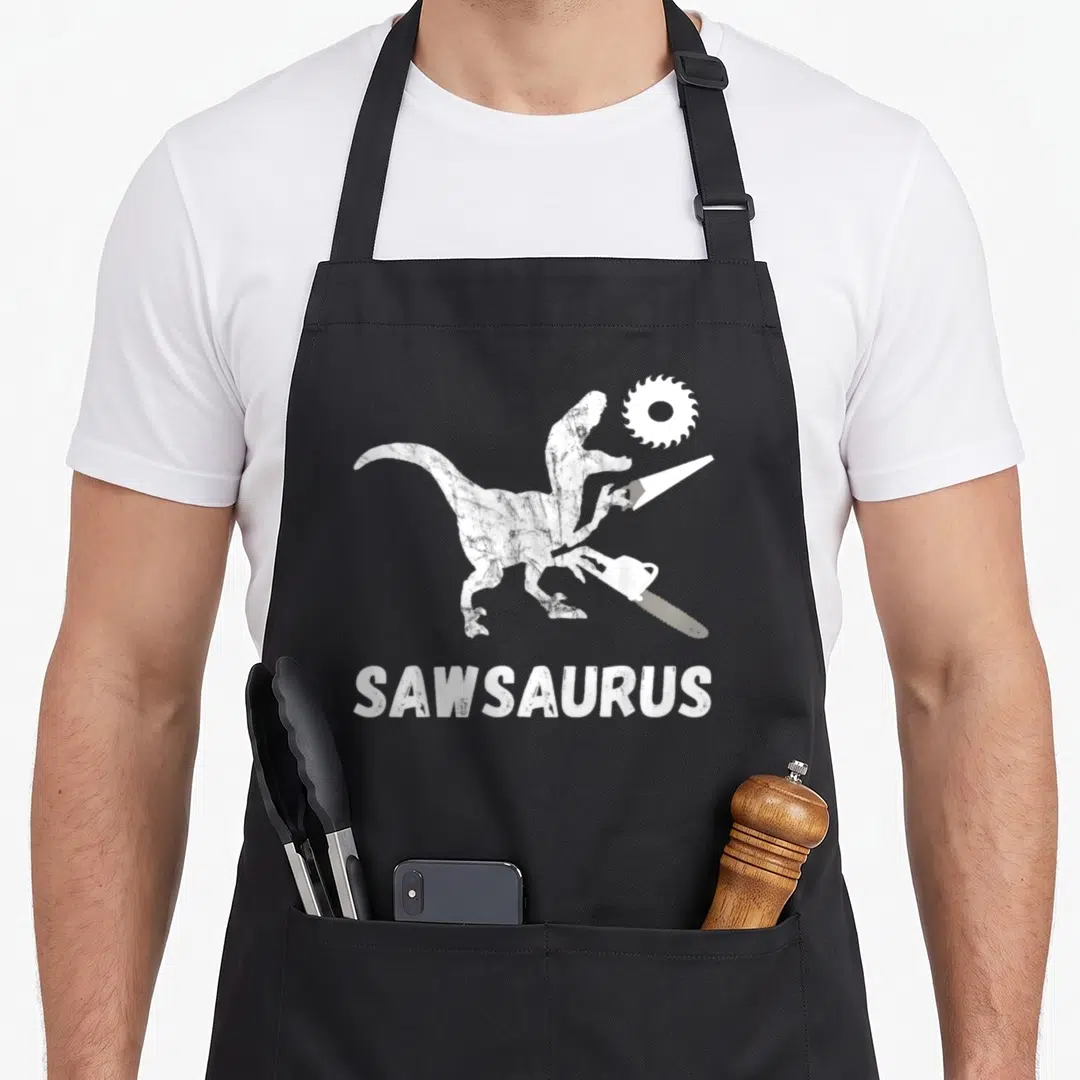 Dinosaur carpentry apron - White cartoon dinosaur holding chainsaw and hand saw with Sawsaurus text below — shown on a black cotton canvas apron against a white background
