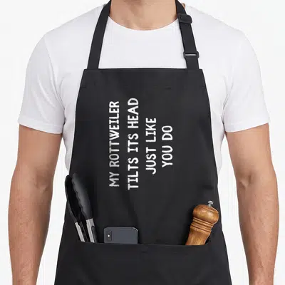 Funny rottweiler dog apron - White vertical text reading MY ROTTWEILER TILTS ITS HEAD JUST LIKE YOU DO on black fabric — shown on a black cotton canvas apron against a white background