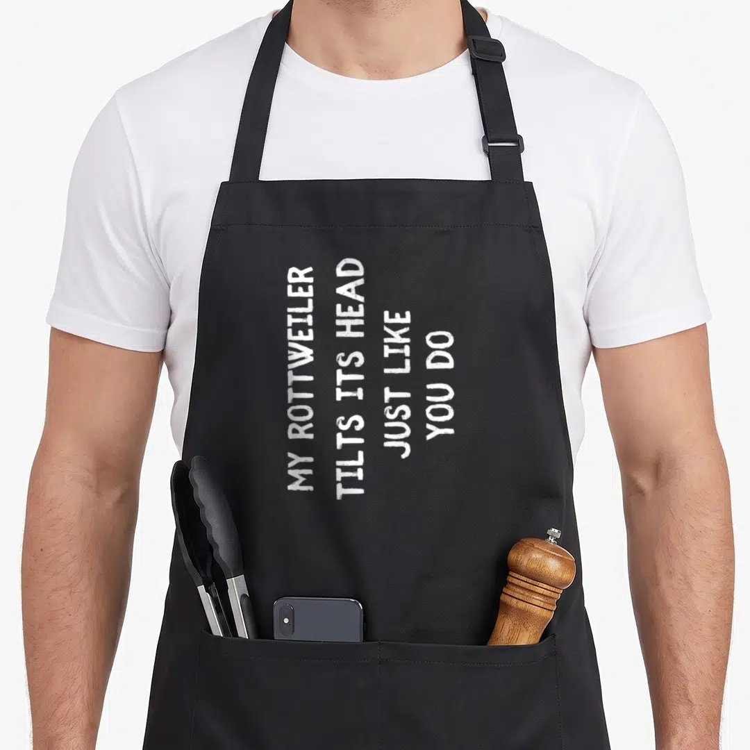 Funny rottweiler dog apron - White vertical text reading MY ROTTWEILER TILTS ITS HEAD JUST LIKE YOU DO on black fabric — shown on a black cotton canvas apron against a white background