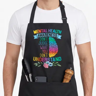 Judge depression awareness apron - Rainbow gradient brain with mental health text and pink blue flowers at bottom — shown on a black cotton canvas apron against a white background