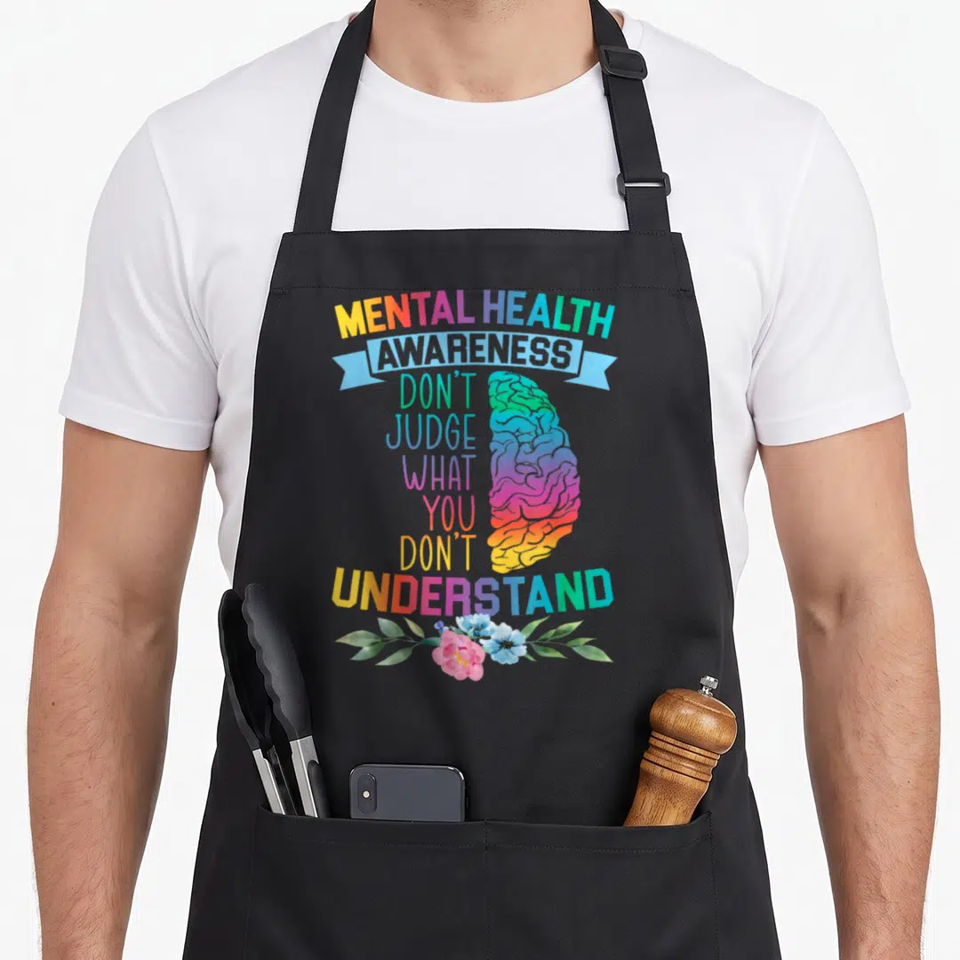 Judge depression awareness apron - Rainbow gradient brain with mental health text and pink blue flowers at bottom — shown on a black cotton canvas apron against a white background