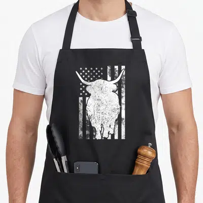 Highland american flag apron - Distressed white highland cow silhouette over stars and stripes flag pattern — shown on a black cotton canvas apron against a white background