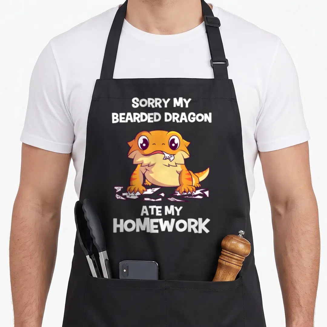 Sorry bearded dragon apron - Cartoon orange bearded dragon chewing paper with SORRY MY BEARDED DRAGON ATE MY HOMEWORK — shown on a black cotton canvas apron against a white background
