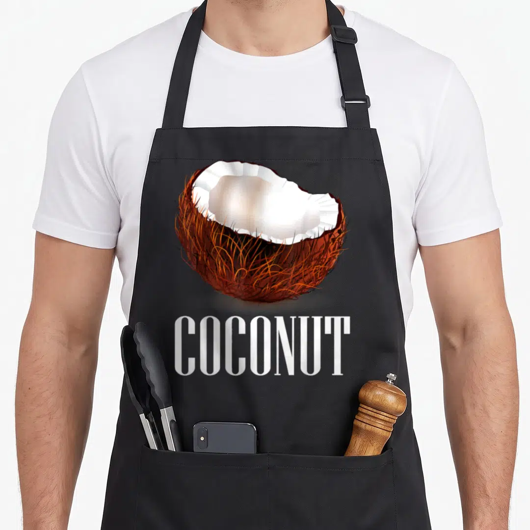 Cute coconut apron - Realistic halved brown coconut with white meat above bold white COCONUT text — shown on a black cotton canvas apron against a white background