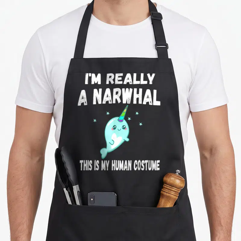 Costume narwhal apron - Light blue cartoon narwhal with rainbow tusk and text I'm Really a Narwhal This Is My Human — shown on a black cotton canvas apron against a white background