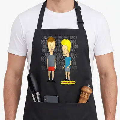 Laughing beaverton apron - Beavis and Butt-Head standing before HUH-HUH-HUH text with yellow title burst — shown on a black cotton canvas apron against a white background