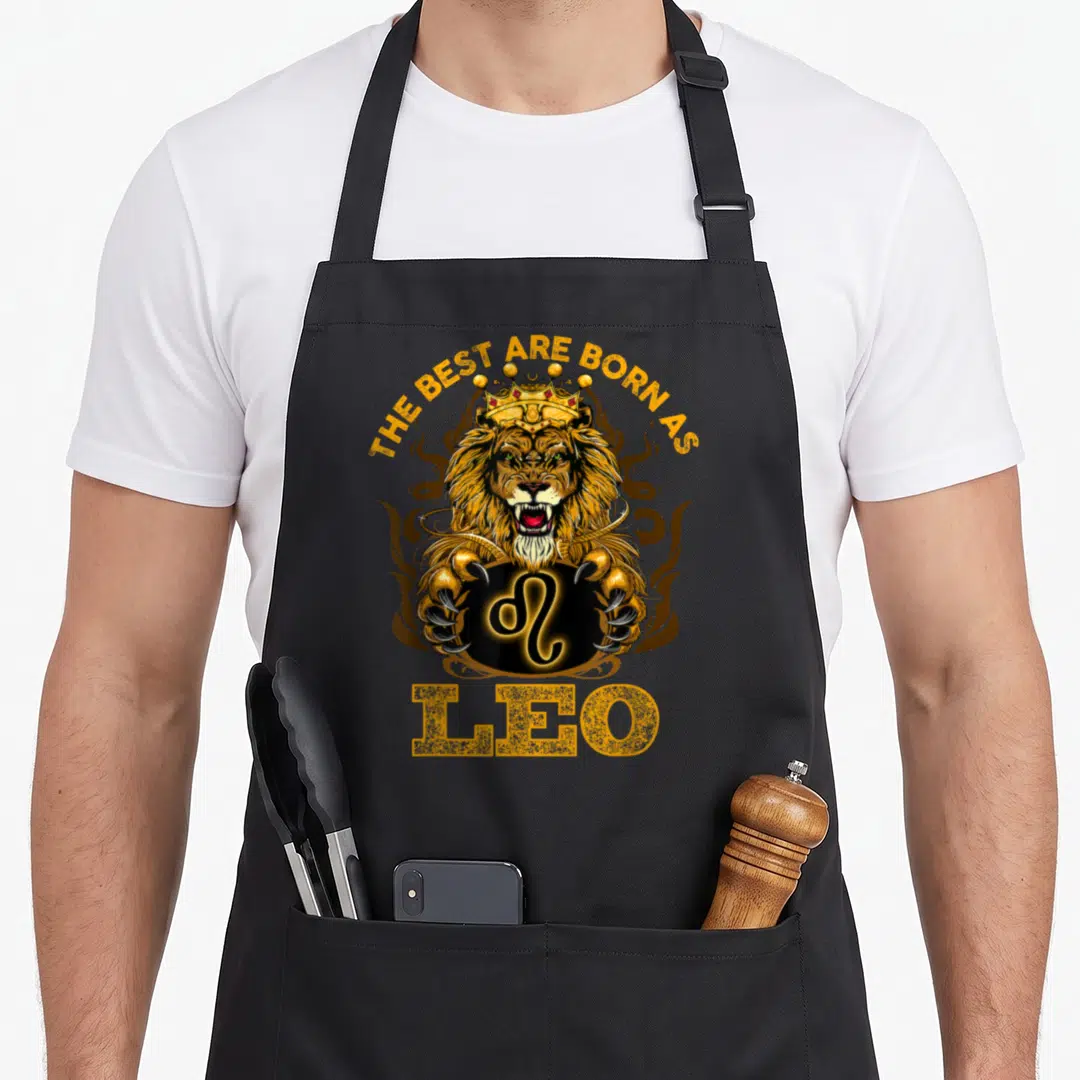 Lion july birthday apron - Realistic crowned lion holding Leo zodiac symbol with gold and brown text — shown on a black cotton canvas apron against a white background
