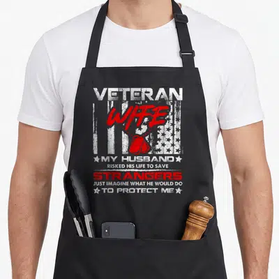 Wife military best husband apron - Distressed white and red text with dog tags and USA flag reading VETERAN WIFE MY — shown on a black cotton canvas apron against a white background