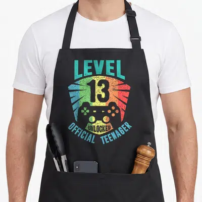 Cute unlocked birthday apron - Retro game controller with Level 13 Unlocked Official Teenager text on striped background — shown on a black cotton canvas apron against a white background