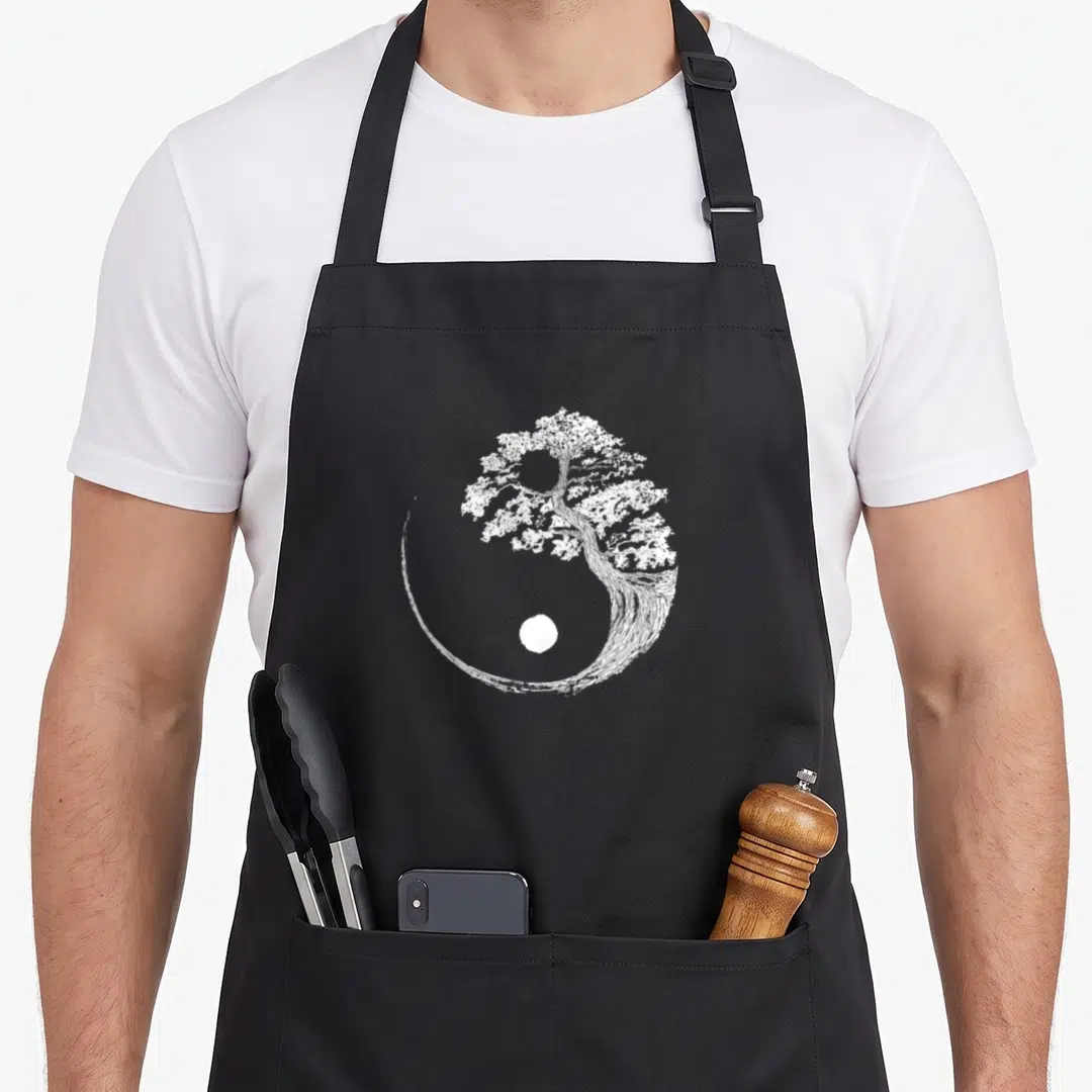 Japanese buddhist tree apron - White line-art bonsai tree forming yin yang shape with dot on black background — shown on a black cotton canvas apron against a white background