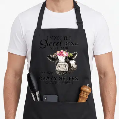 Sweet door apron - Cartoon cow head with pink flower crown and black text about being a crazy heifer down the street — shown on a black cotton canvas apron against a white background