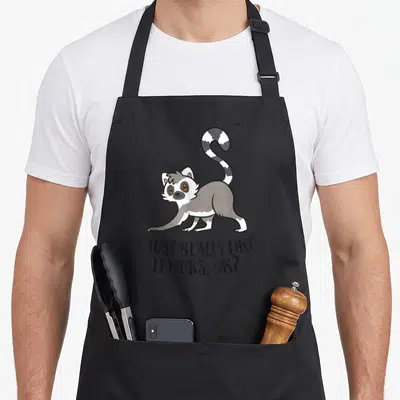 Cute lemur apron - Cartoon lemur with striped tail above text I JUST REALLY LIKE LEMURS OK — shown on a black cotton canvas apron against a white background