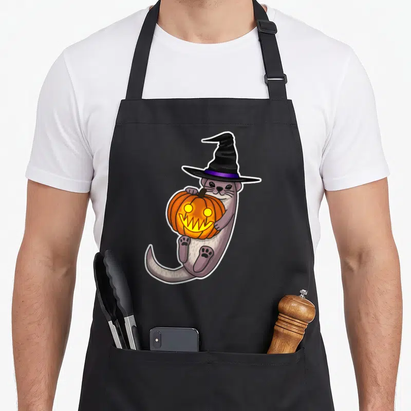 Otter halloween apron - Cartoon grey otter in black witch hat holding orange jack-o-lantern pumpkin — shown on a black cotton canvas apron against a white background