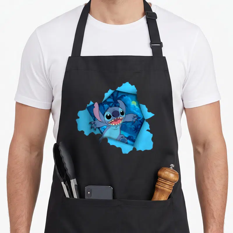 Tropical novelty apron - Stitch bursts through jagged light blue paper revealing a dark blue tropical night scene — shown on a black cotton canvas apron against a white background