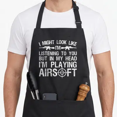 Airsoft gun shooting apron - White distressed text with two rifles and crosshair symbol on dark background — shown on a black cotton canvas apron against a white background