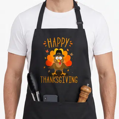 Family thanksgiving day aprons - Cartoon turkey wearing a black hat between HAPPY and THANKSGIVING text with orange — shown on a black cotton canvas apron against a white background