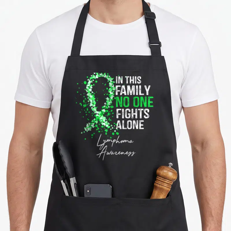 Lymphoma family aprons - Green and white ribbon made of small hearts next to text IN THIS FAMILY NO ONE FIGHTS ALONE — shown on a black cotton canvas apron against a white background