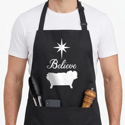Believe baby christmas apron - White star above Believe script and manger silhouette on grey background — shown on a black cotton canvas apron against a white background