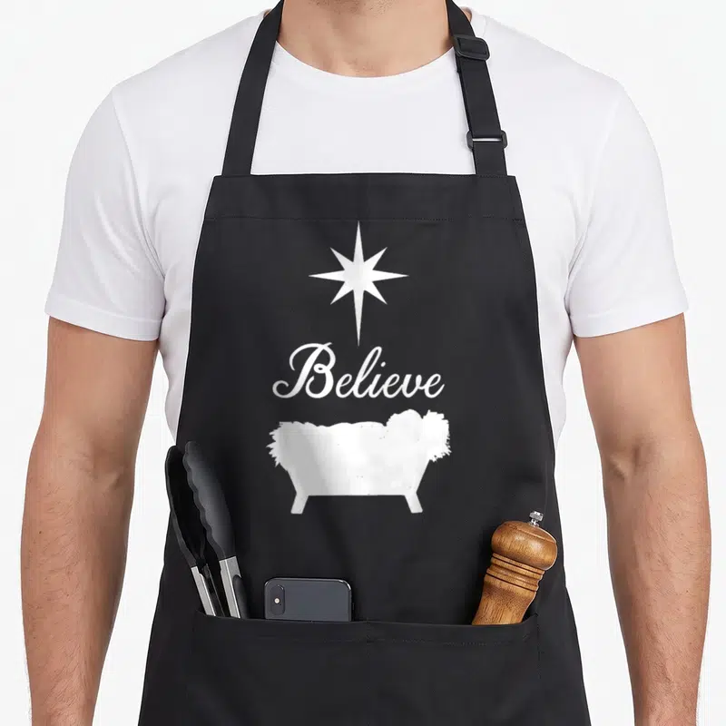Believe baby christmas apron - White star above Believe script and manger silhouette on grey background — shown on a black cotton canvas apron against a white background