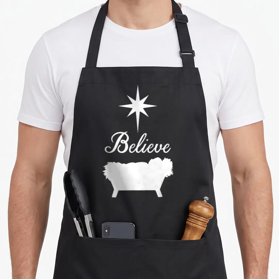 Believe baby christmas apron - White star above Believe script and manger silhouette on grey background — shown on a black cotton canvas apron against a white background