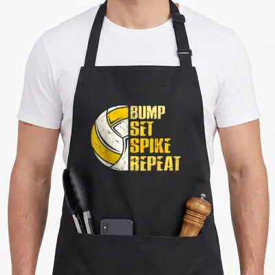 Spike bump apron - Yellow and white volleyball next to stacked distressed text reading BUMP SET SPIKE REPEAT — shown on a black cotton canvas apron against a white background
