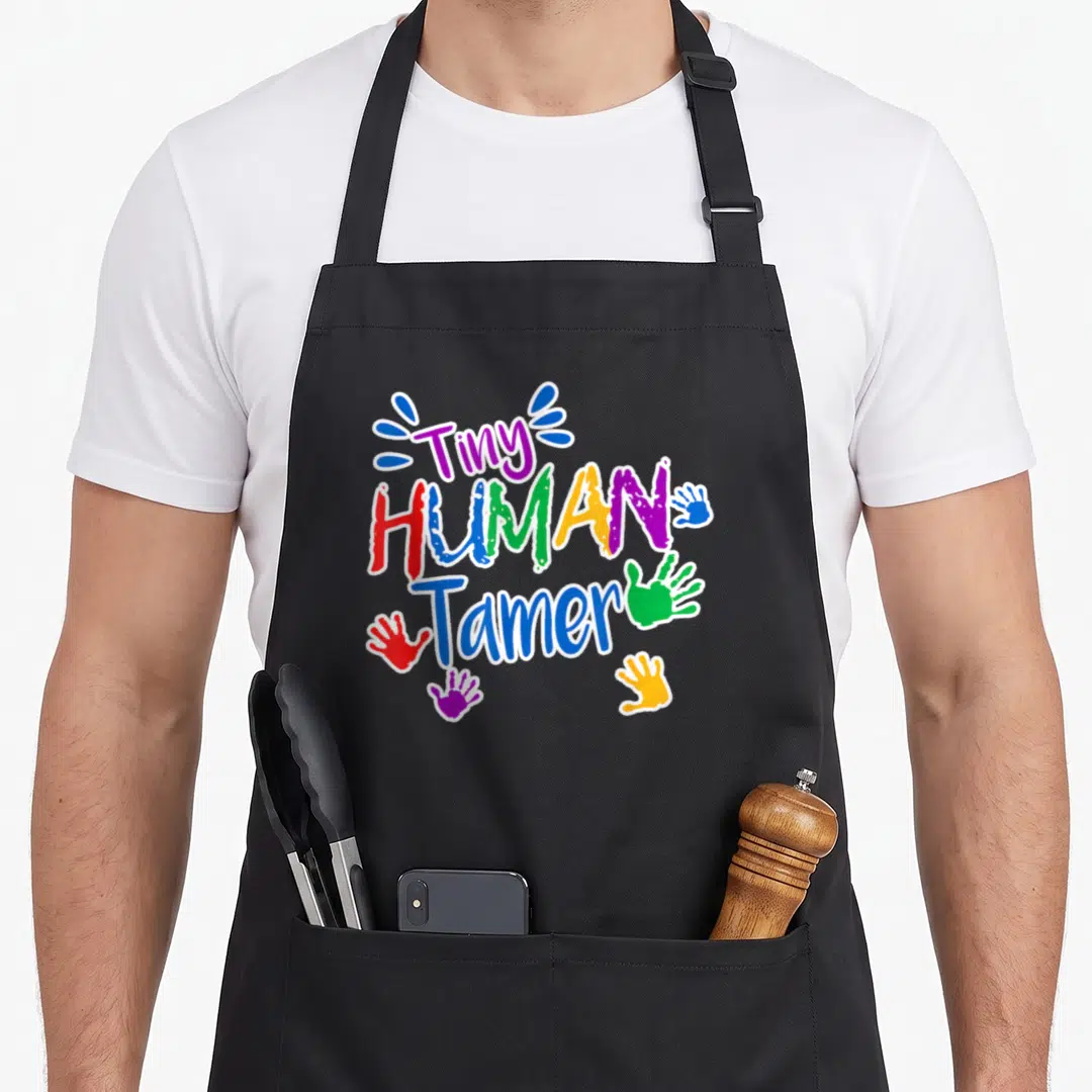 Daycare tiny chef apron - Tiny HUMAN Tamer text with colorful handprints and blue splatters — shown on a black cotton canvas apron against a white background