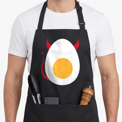 Boiled egg apron - White fried egg with yellow yolk, red horns, and red devil tail — shown on a black cotton canvas apron against a white background