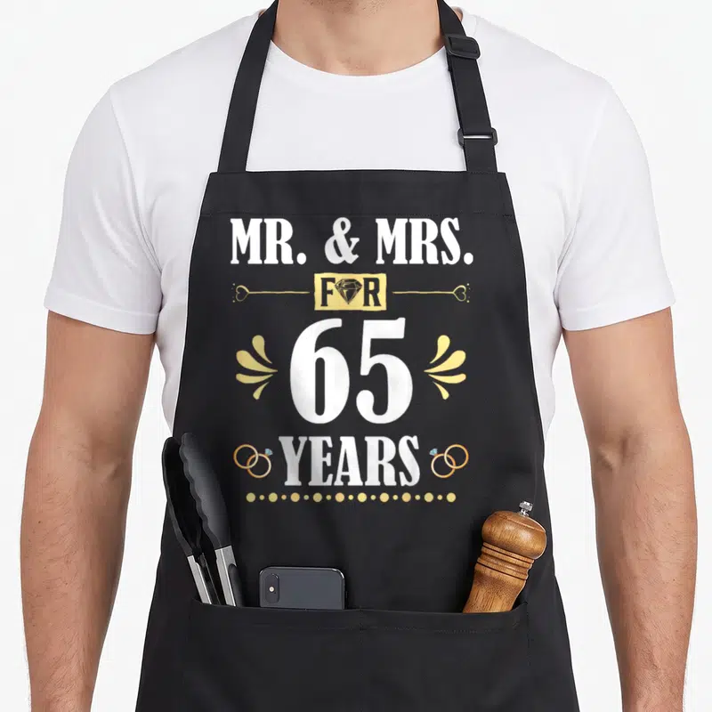 65th mr mrs aprons - Stacked white and gold text MR MRS FOR 65 YEARS with diamond and ring graphics — shown on a black cotton canvas apron against a white background