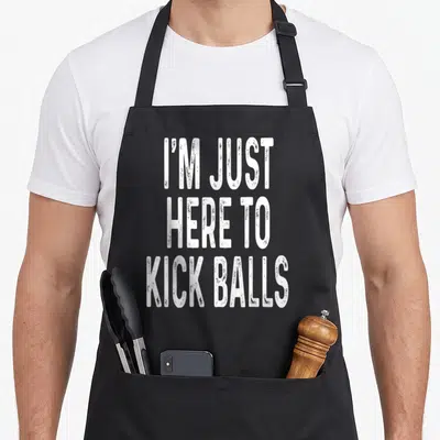 Kickball game aprons - White stacked text reading I'M JUST HERE TO KICK BALLS on black fabric — shown on a black cotton canvas apron against a white background