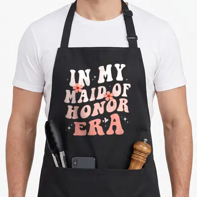 Honor bridesmaid aprons - Retro text reading In My Maid of Honor Era with pink flowers and sparkles — shown on a black cotton canvas apron against a white background