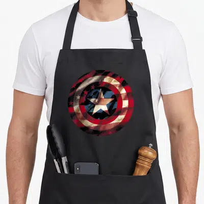 Comic captain apron - Distressed circular shield with red white blue rings and central star — shown on a black cotton canvas apron against a white background