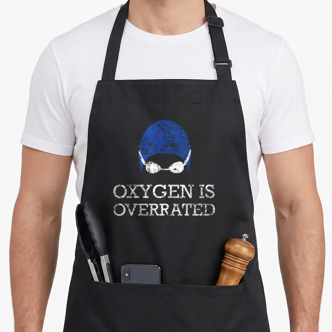 Oxygen swimmer apron - Blue swim cap and white goggles above stacked text Oxygen Is Overrated in distressed style — shown on a black cotton canvas apron against a white background