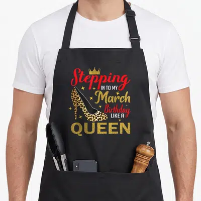 Stepping leopard apron - High heel shoe with Stepping In To My March Birthday Like A Queen text and crown — shown on a black cotton canvas apron against a white background