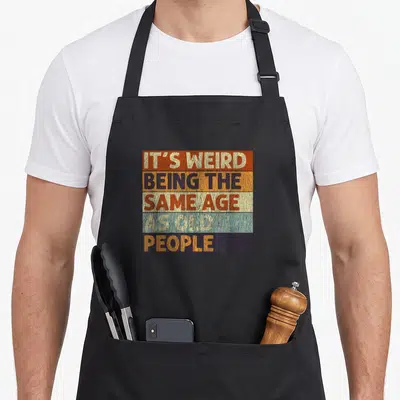 General weird aprons - Retro striped typography reading It's Weird Being the Same Age As Old People in orange blue — shown on a black cotton canvas apron against a white background