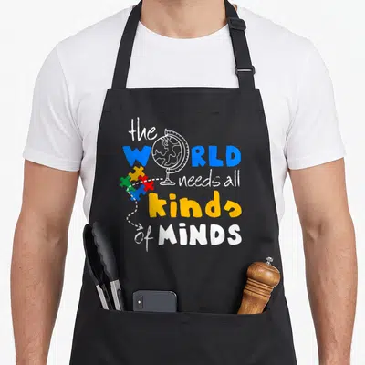 Awareness novelty apron - Globe replacing O in WORLD with blue yellow red green puzzle pieces and dotted arrow — shown on a black cotton canvas apron against a white background