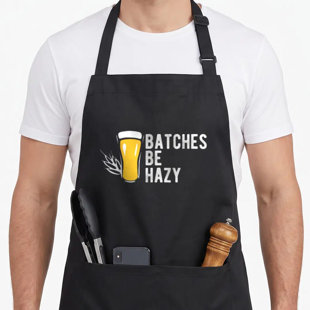 Craft beer brewing apron - Cartoon yellow beer glass with white wheat stalks and stacked text Batches Be Hazy — shown on a black cotton canvas apron against a white background