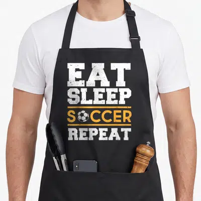 Soccer sleep apron - Stacked white and orange text EAT SLEEP SOCCER REPEAT with ball graphic replacing the O letter — shown on a black cotton canvas apron against a white background