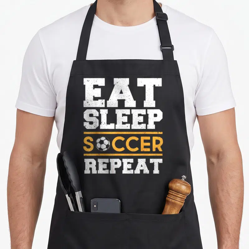 Soccer sleep apron - Stacked white and orange text EAT SLEEP SOCCER REPEAT with ball graphic replacing the O letter — shown on a black cotton canvas apron against a white background