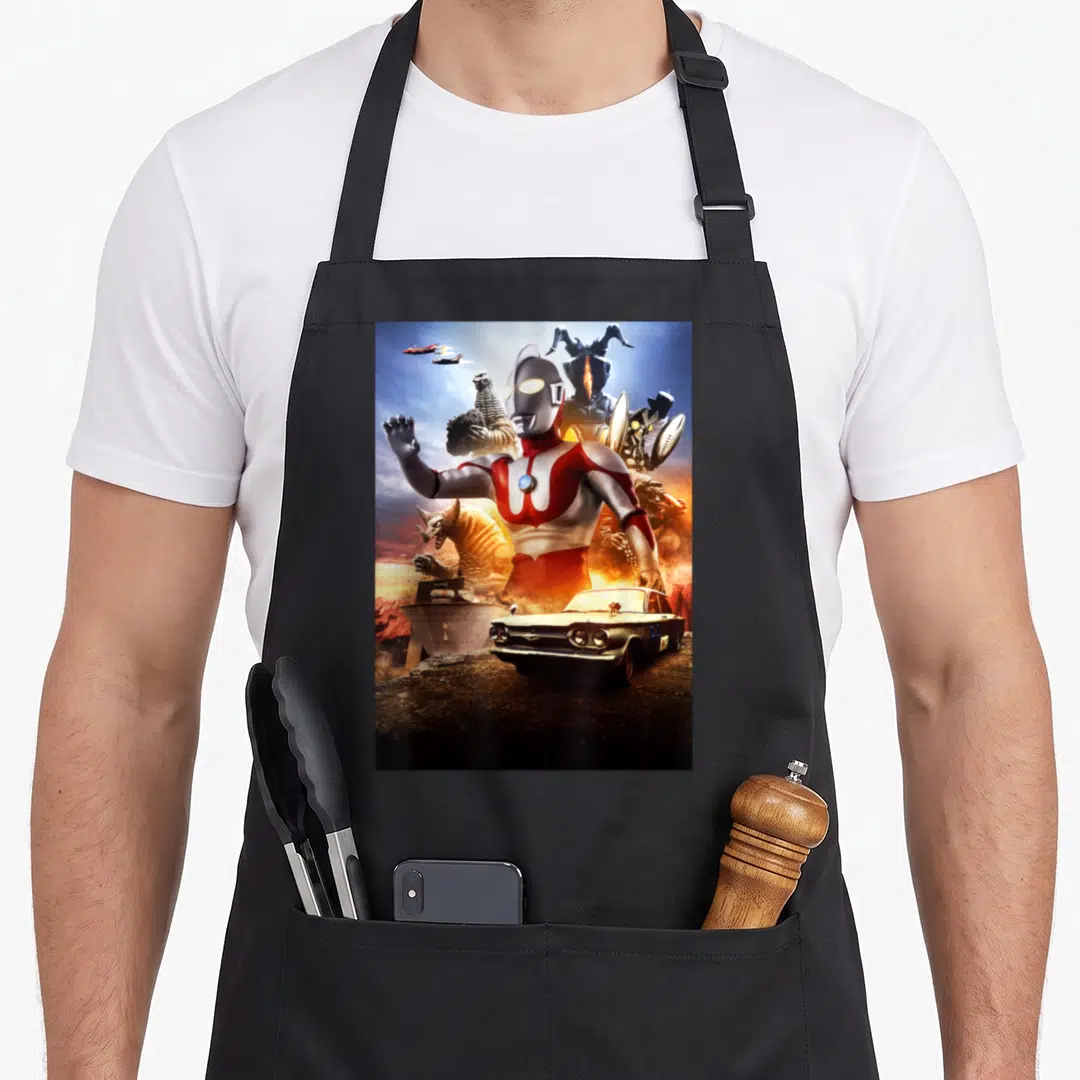 Ultraman b apron - Ultraman central figure surrounded by monsters, vintage car, and flying jets in red silver blue — shown on a black cotton canvas apron against a white background