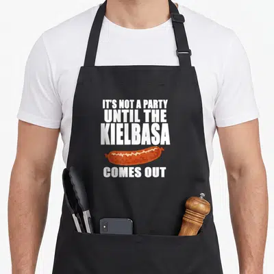 Kielbasa polish aprons - White stacked text reading IT'S NOT A PARTY UNTIL THE KIELBASA COMES OUT above a brown sausage — shown on a black cotton canvas apron against a white background