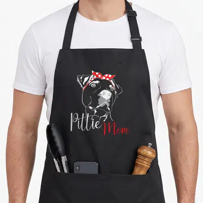Pittie mother's day aprons - Grey line art dog head with red polka dot bandana and Pittie Mom script text — shown on a black cotton canvas apron against a white background
