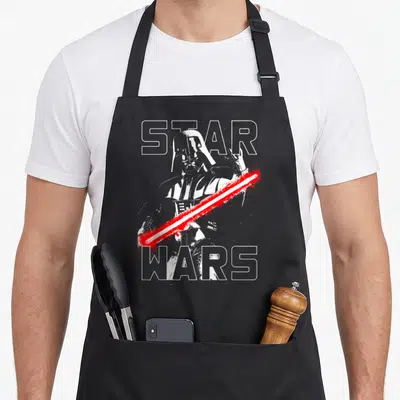 Taunting general apron - Darth Vader behind STAR WARS text with red lightsaber slash in distressed white and grey — shown on a black cotton canvas apron against a white background