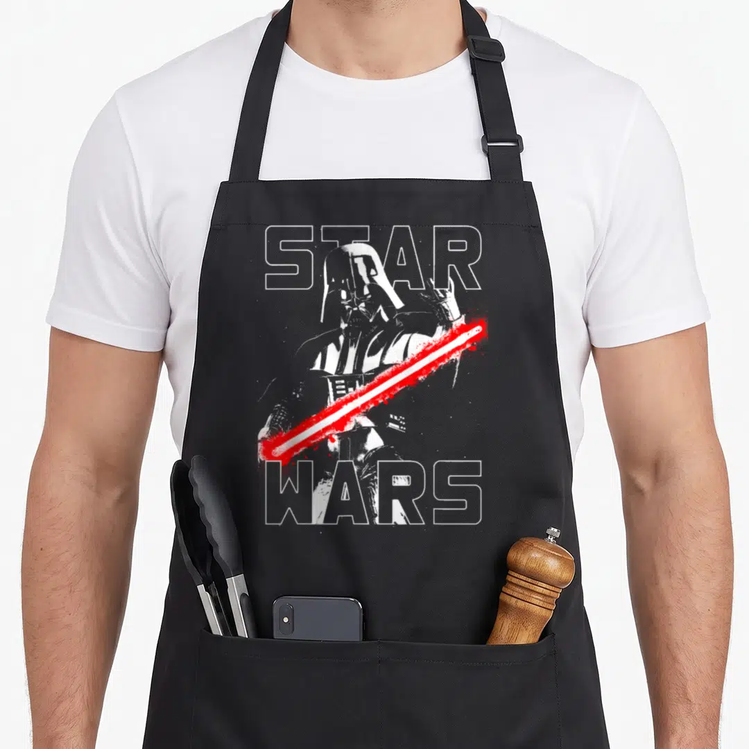 Taunting general apron - Darth Vader behind STAR WARS text with red lightsaber slash in distressed white and grey — shown on a black cotton canvas apron against a white background