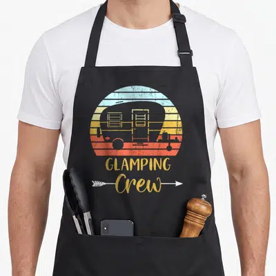 Camping family aprons - Retro camper silhouette over striped sunset with GLAMPING Crew text and white arrow — shown on a black cotton canvas apron against a white background
