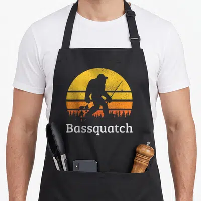Sasquatch fishing aprons - Grey Bigfoot silhouette fishing in front of yellow orange sun with Bassquatch text — shown on a black cotton canvas apron against a white background