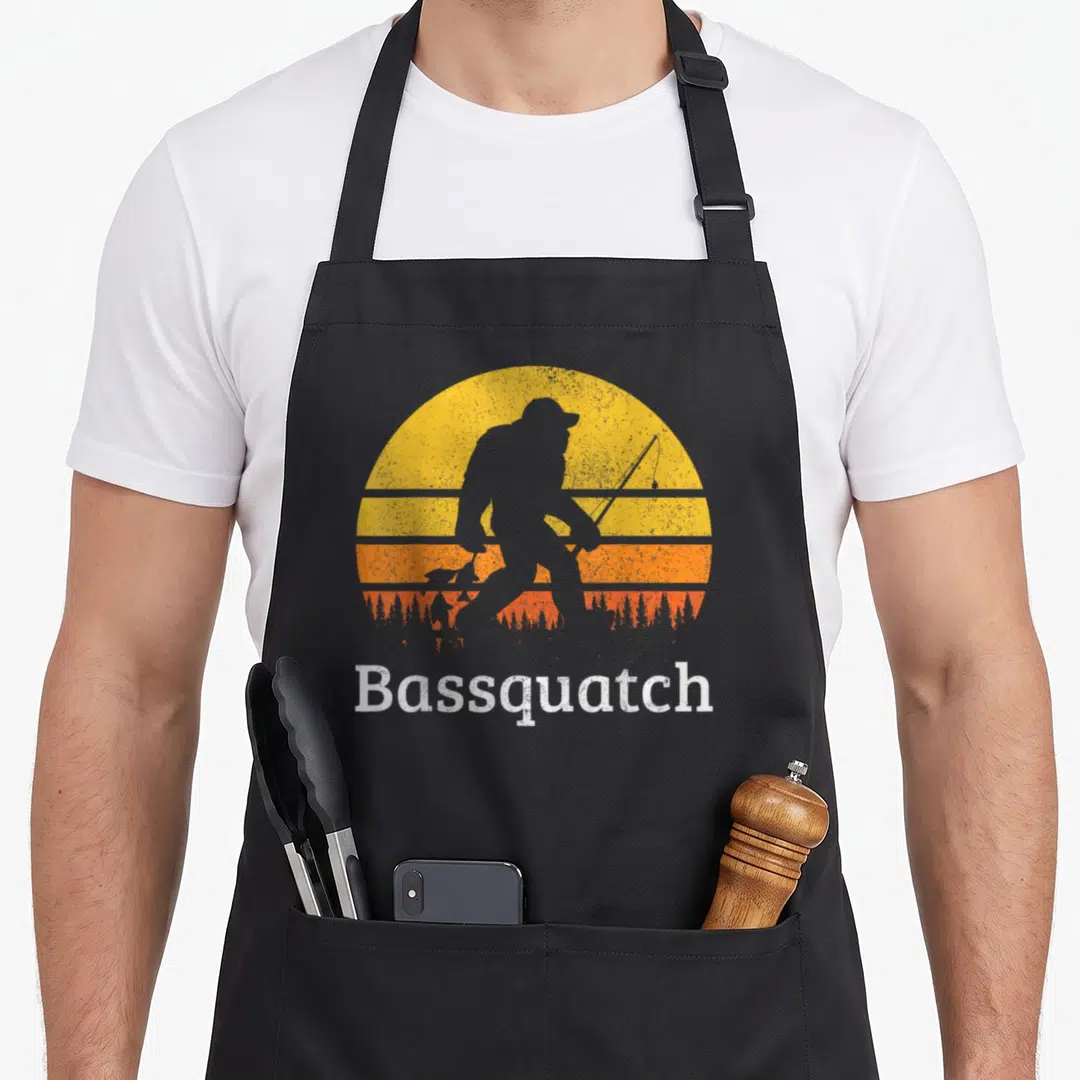 Sasquatch fishing aprons - Grey Bigfoot silhouette fishing in front of yellow orange sun with Bassquatch text — shown on a black cotton canvas apron against a white background
